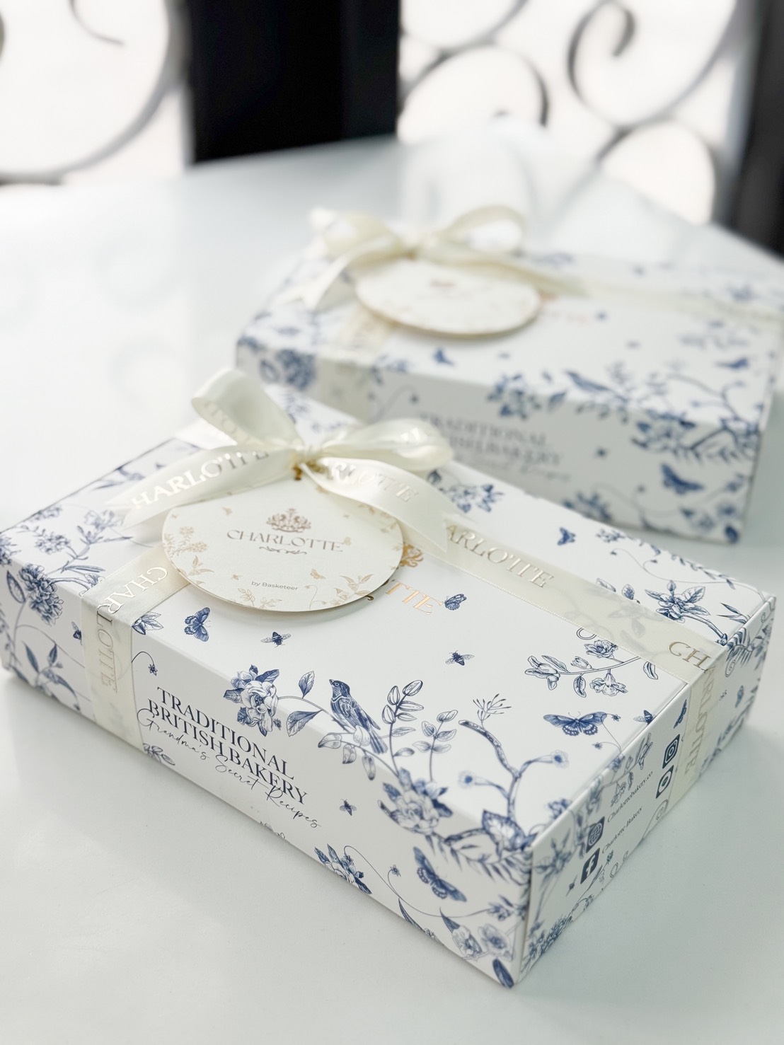 Two elegantly wrapped bakery boxes with blue floral designs and white ribbons sit on a white surface. Each box has a round gift tag and the words 