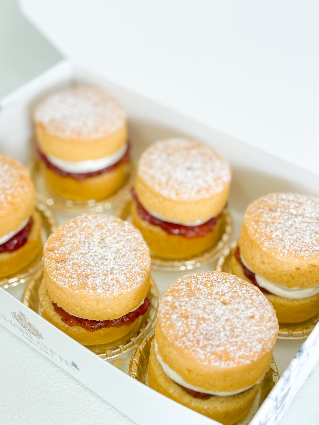 Six small sponge cakes filled with cream and strawberry jam, topped with icing sugar, arranged neatly in a white box.