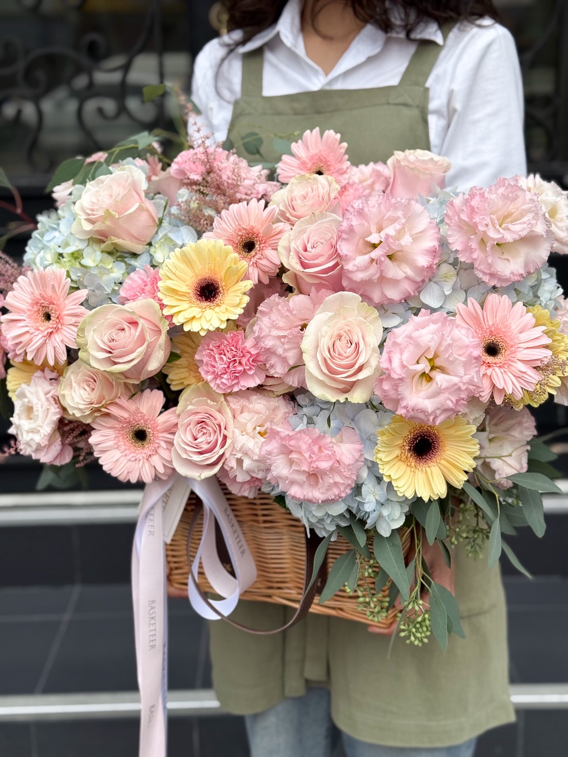 A person in a green apron and white shirt holds a large Carnations & Gerbera Basket Bouquet filled with pink roses, carnations, and yellow blooms tied with ribbon. The background is blurred and dark.