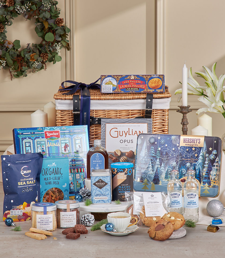 A full winter-themed gourmet hamper with a wicker basket filled with blue-packaged snacks, pasta, cookies, chocolates, tea, and coffee arranged with silver ornaments and snow-inspired décor