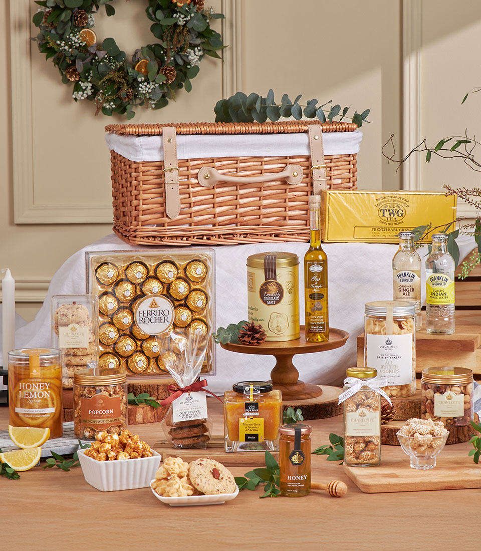 The Golden Breakfast & Picnic Gift Basket features a festive wicker hamper filled with Ferrero Rocher chocolates, honey, biscuits, popcorn, lemon tea, pasta, olive oil, and drinks—all beautifully arranged on a white tablecloth.