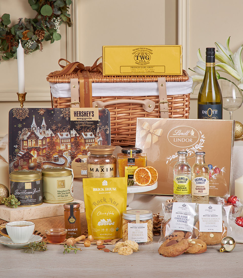 A Luxury Gourmet Celebration Hamper contains wine, chocolates, biscuits, honey, tea, marmalade, dried fruit, nuts, and preserves—elegantly arranged in front of a wicker basket with candles and holiday decorations in the background.