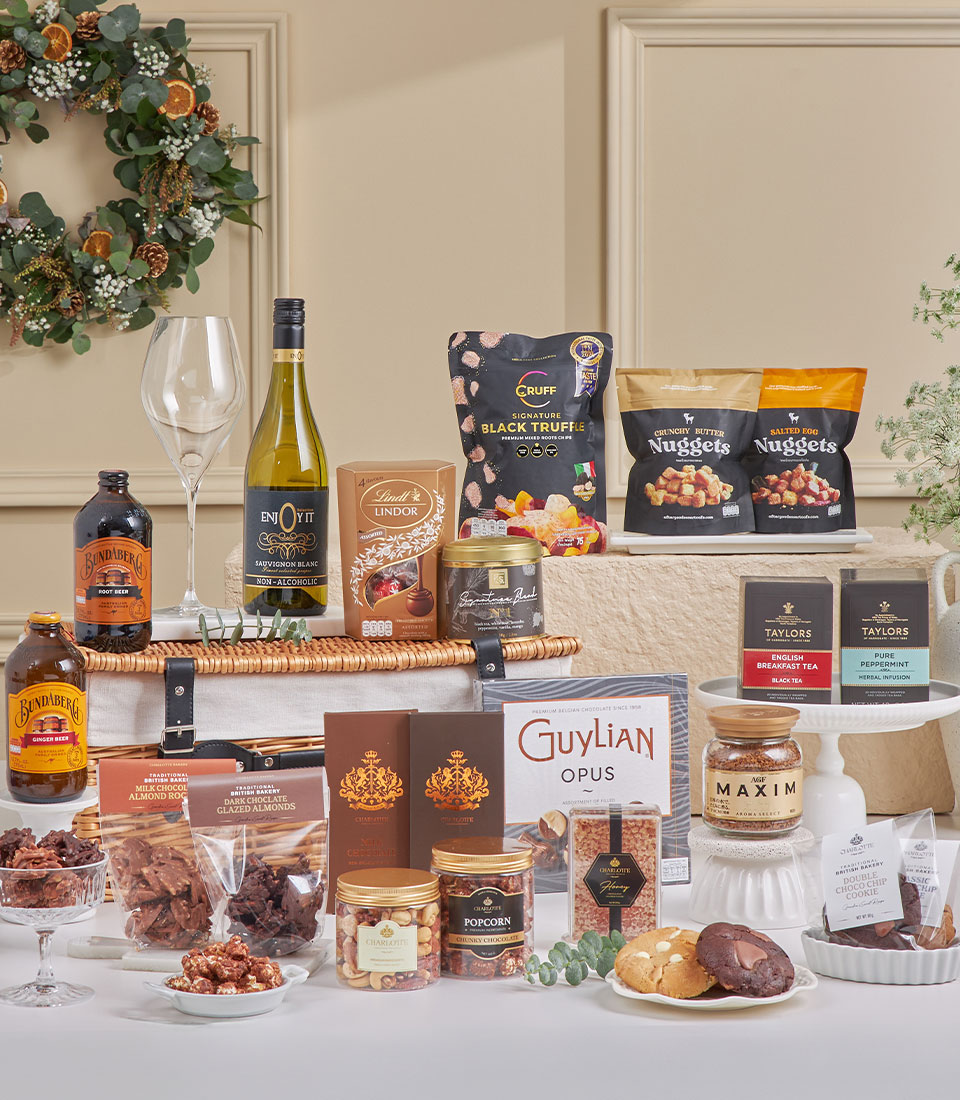 The Ultimate Chocolate & Tea Celebration Hamper features wine, chocolates, biscuits, popcorn, truffle crisps, fudge, coffee, maple syrup, and snacks—artfully arranged with two wine glasses and a festive wreath in the background.