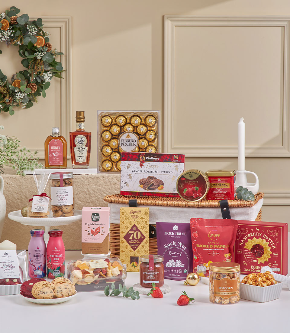 A festive Prosperity New Year gift hamper arranged with chocolates, biscuits, popcorn, preserves, drinks, and gourmet treats graces the table. Decorative greenery, a candle, a wall wreath, and strawberries complete this elegant gourmet display.
