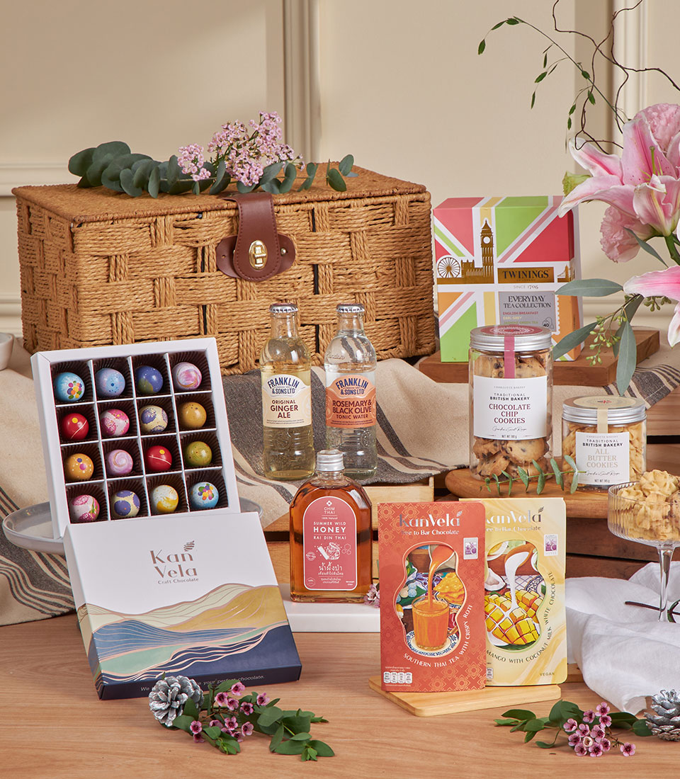 A wicker picnic basket with a leather strap is surrounded by assorted gourmet treats, including the London & Thai Tea Chocolate Hamper, flavoured drinks, biscuits, snacks, and floral decorations on a wooden table.