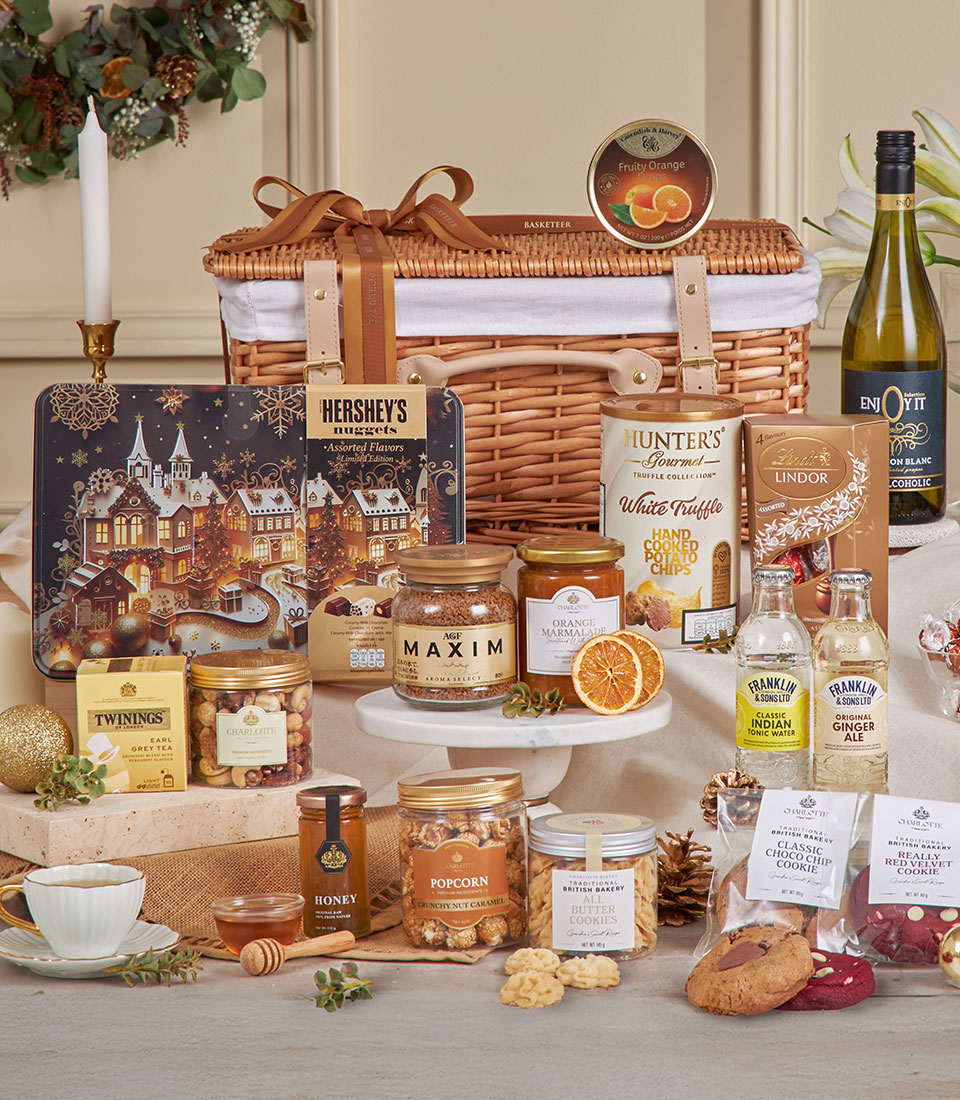 A Premium Golden Gourmet Hamper in a wicker basket brims with festive treats—chocolates, biscuits, popcorn, honey, tea, jam, truffle crisps, gourmet snacks, and a bottle of white wine—beautifully arranged on a table.