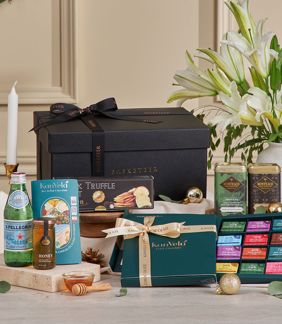 A luxurious Premium Chocolate & Tea Hamper with a black box tied with a ribbon, assorted chocolates, honey, sparkling water, crackers, and gourmet tea, displayed on a table with white lilies and a lit candle in the background.