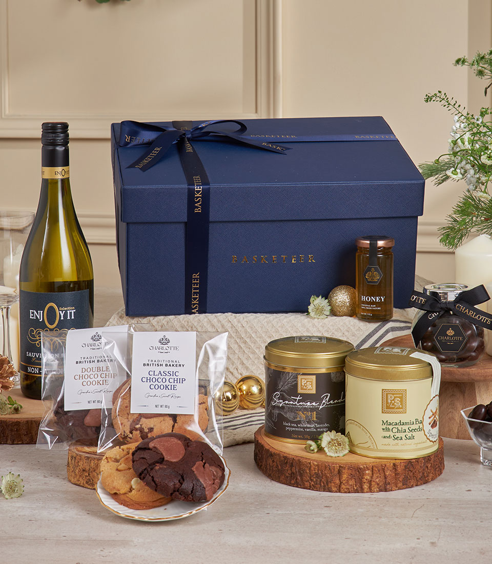 A navy blue Non-Alcoholic White Wine Dessert Gift Box with a ribbon sits beside honey, chocolate chip biscuits, butter biscuits, and jars of nuts and biscuits, all arranged on a wooden table with festive decorations.