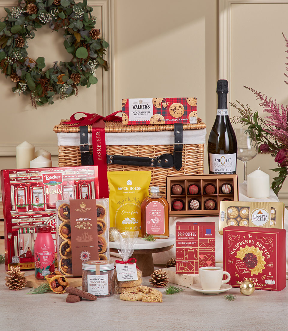 A full Christmas gourmet hamper displayed with a wicker basket filled with red and gold holiday treats including chocolate truffles, honey, cookies, a yellow snack pouch, and a sparkling drink bottle decorated with pinecones and candles