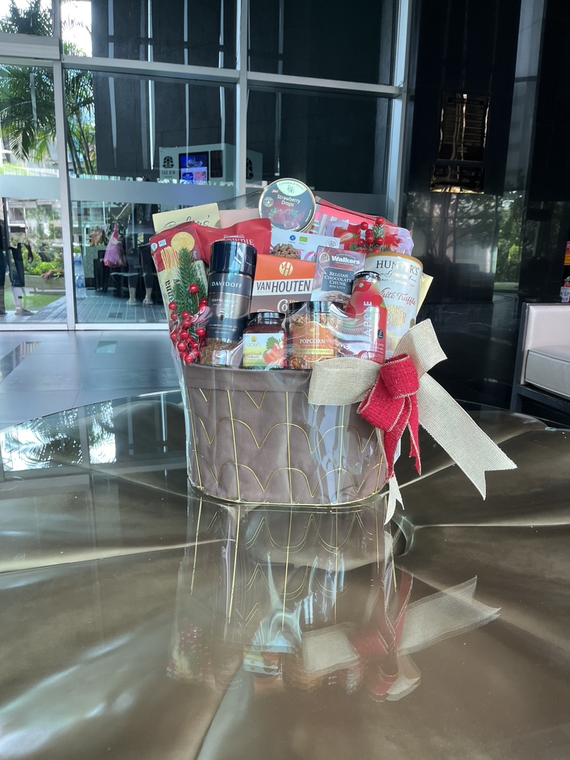 The Deluxe Red Gourmet Snack Hamper, filled with assorted snacks, chocolates, and drinks, is adorned with red berries, greenery, and a gold bow as it sits on a reflective table in a modern building foyer.