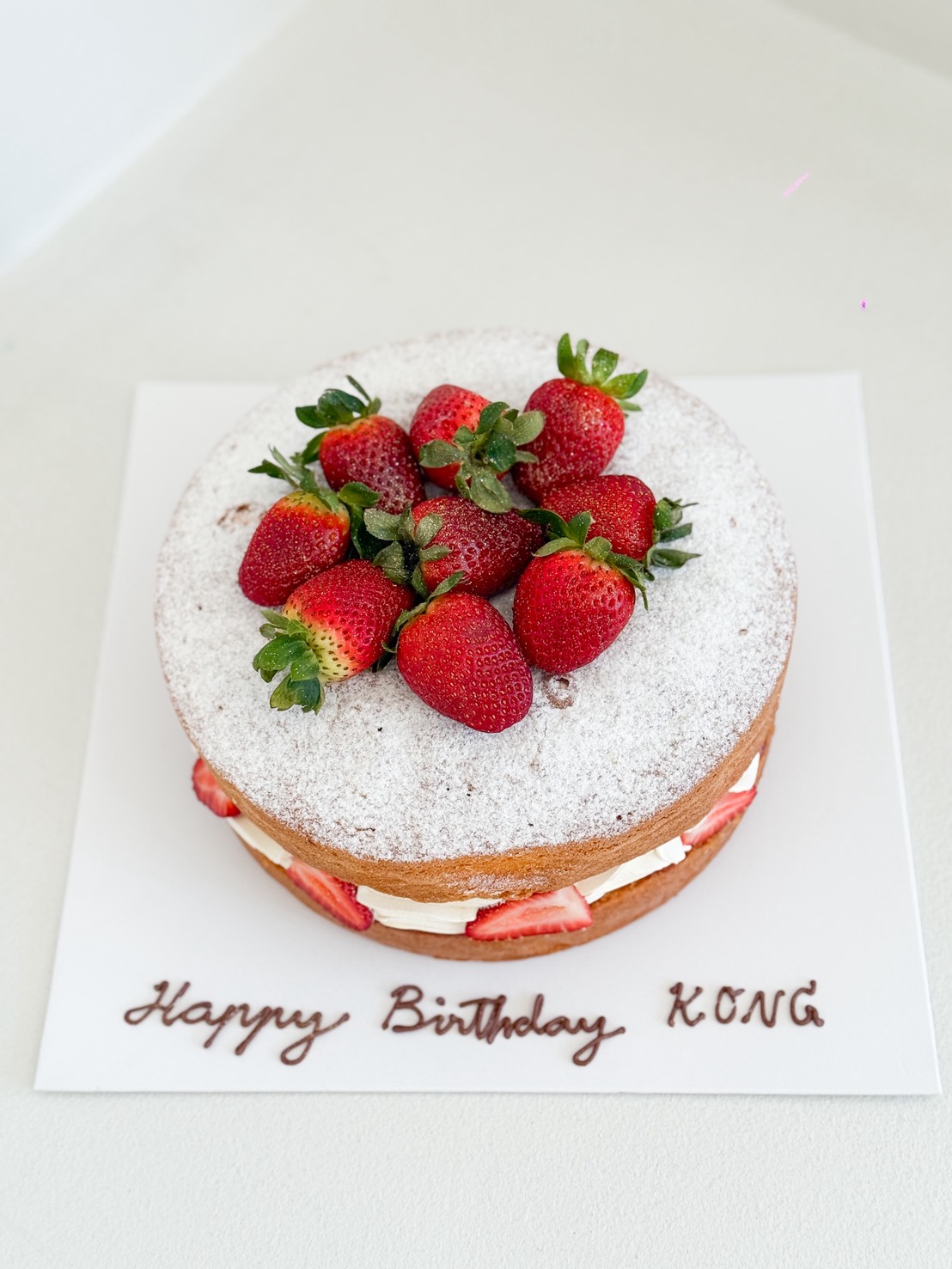 A round cake topped with whole strawberries and icing sugar, with sliced strawberries and cream filling inside. 