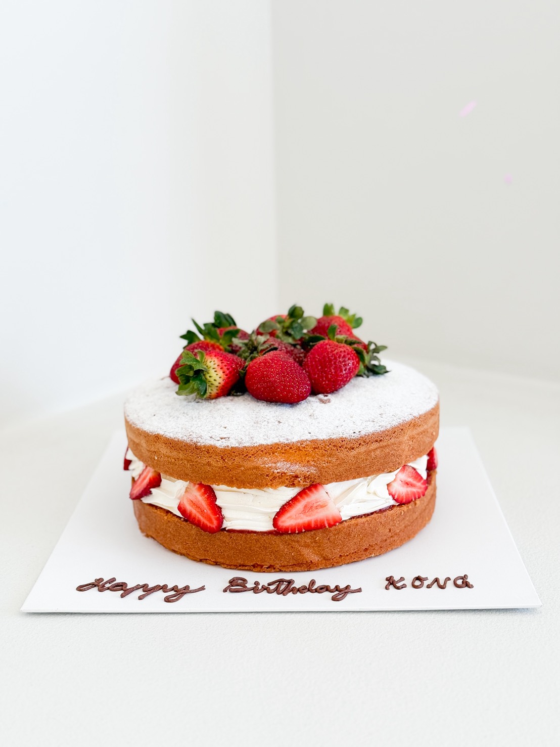 A round sponge cake filled with whipped cream and sliced strawberries, topped with whole strawberries and icing sugar, sits on a white board with 