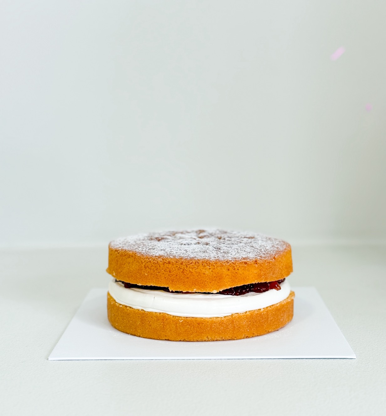 A Victoria sponge cake with two golden layers, filled with cream and jam, topped with a light dusting of icing sugar, sits on a white square board against a plain background.