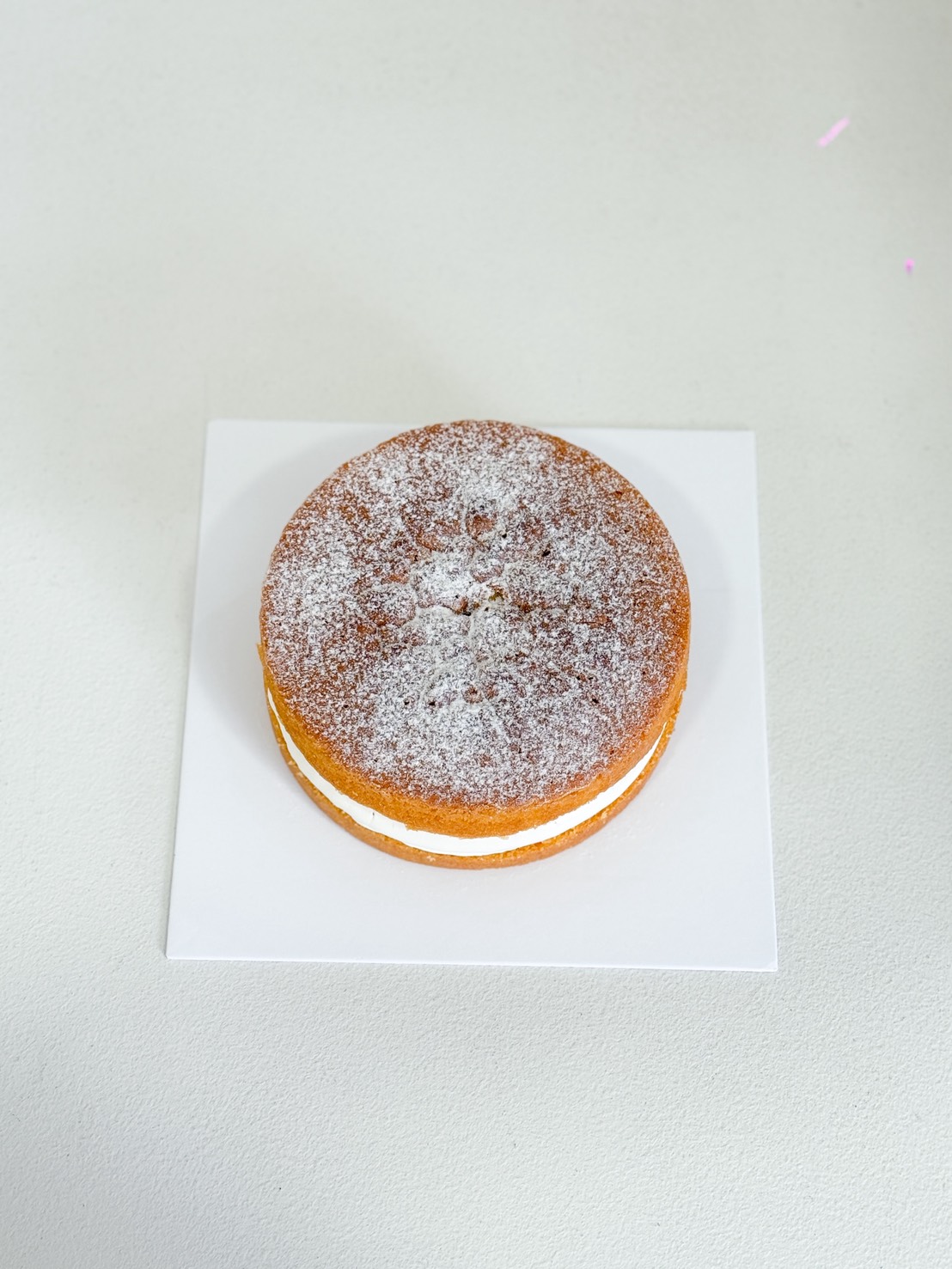 A round, two-layer sponge cake with white cream filling and a dusting of icing sugar on top, placed on a white square board against a light background.