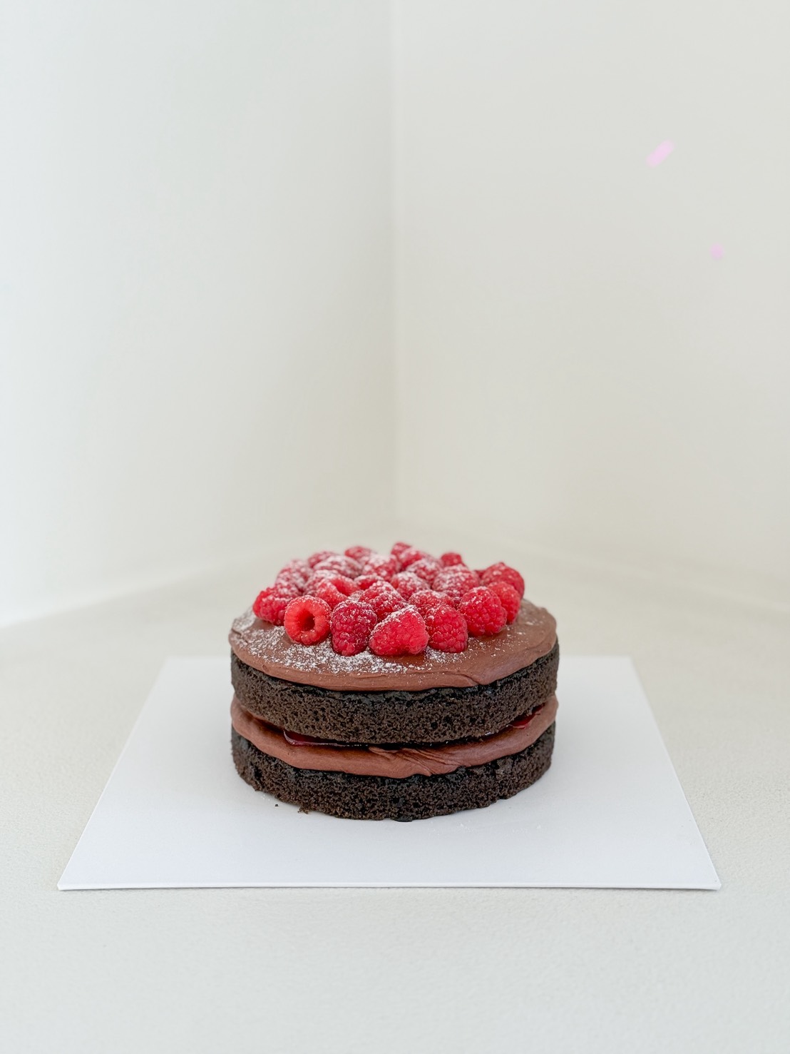 A round chocolate layer cake with chocolate icing and raspberries on top, dusted with icing sugar, sits on a white square board against a light background.