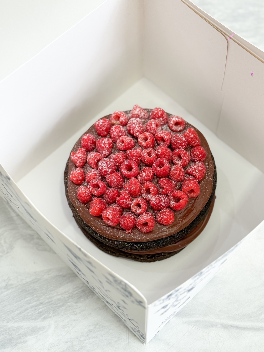 A chocolate layer cake topped with fresh raspberries and a dusting of icing sugar, placed inside a white bakery box on a light-coloured surface.
