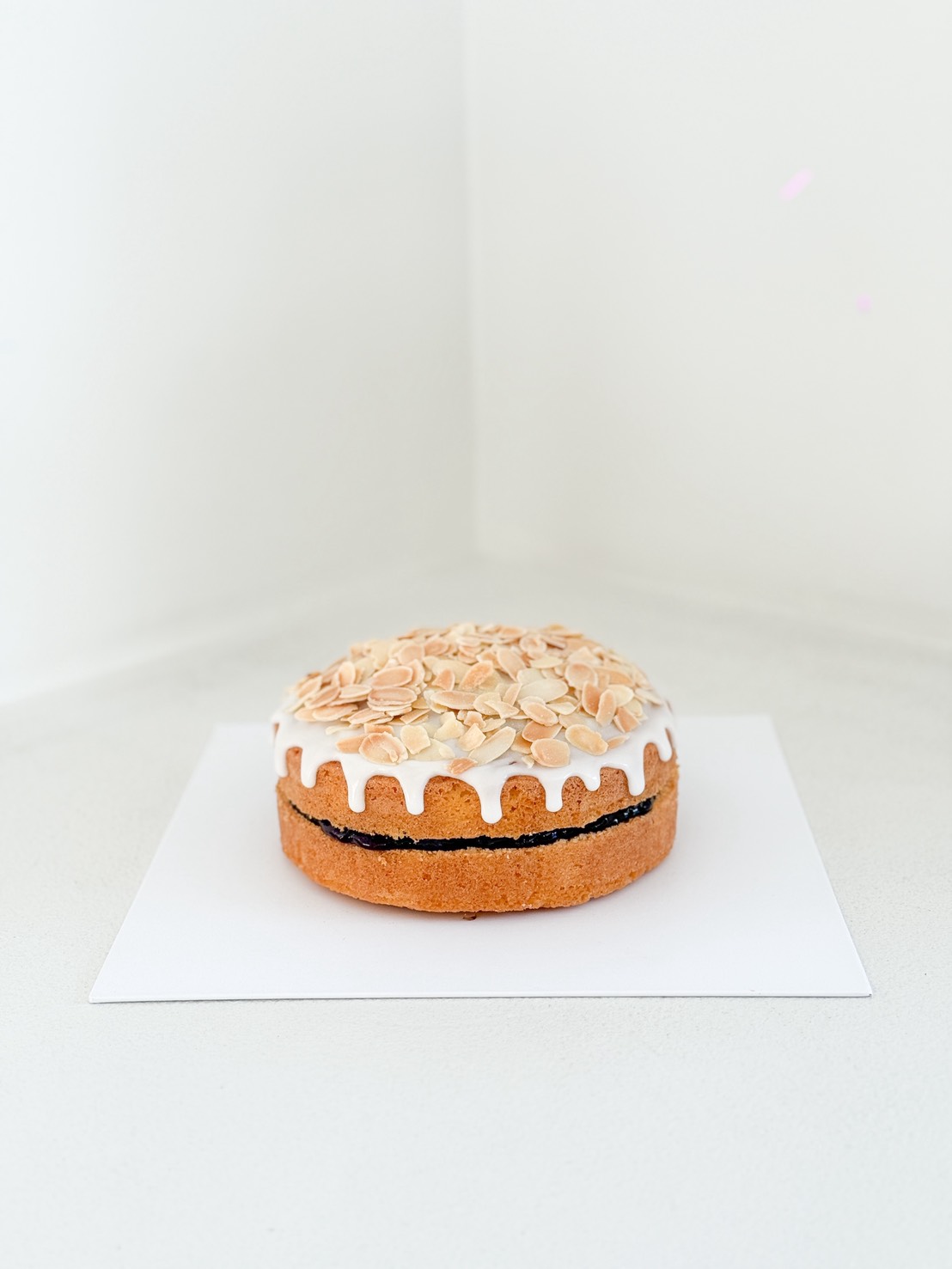 A round cake with white icing dripping down the sides, topped with sliced almonds, and a visible dark filling layer, placed on a white square sheet in a minimalist white setting.