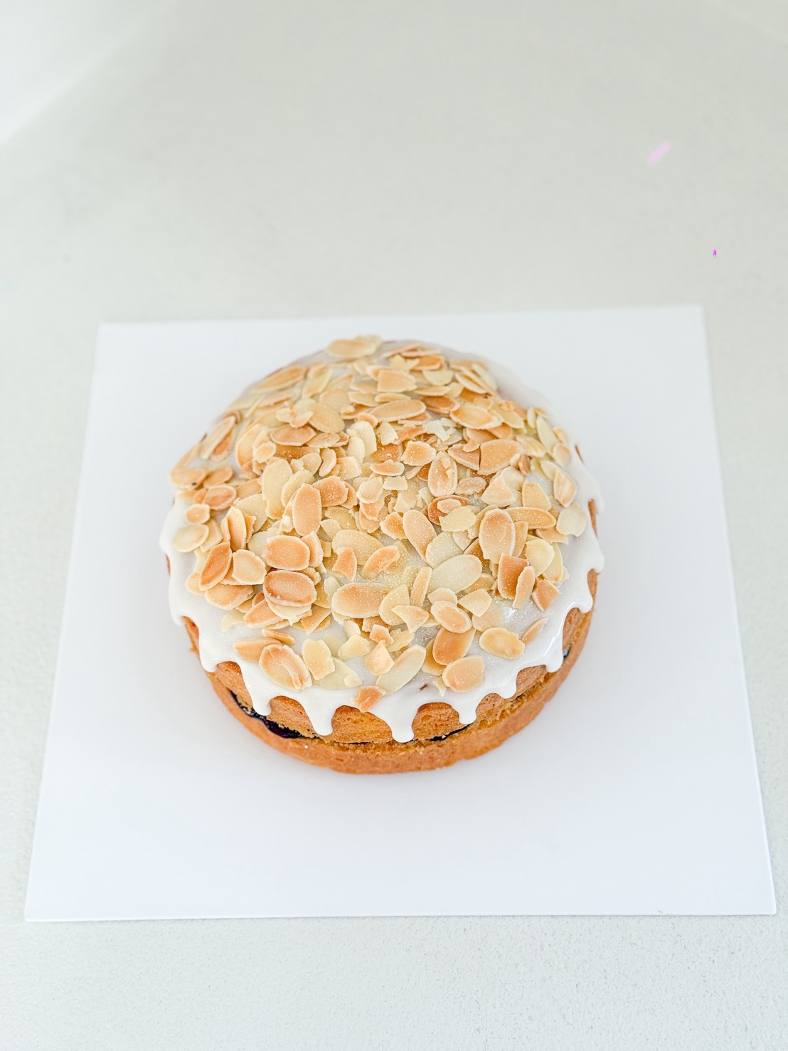 A round cake topped with white icing and a generous layer of sliced, toasted almonds, placed on a white square sheet against a light background.