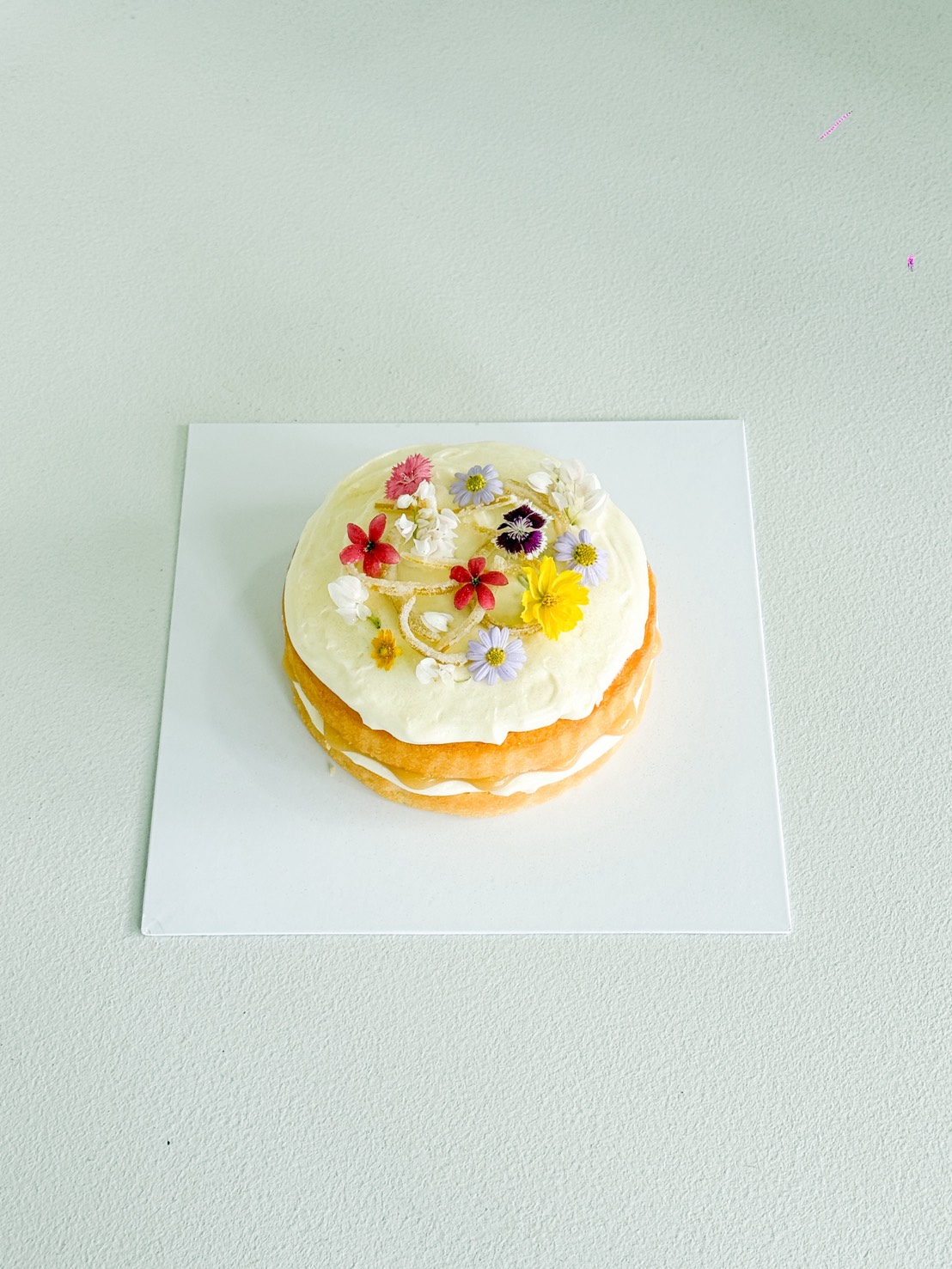 A round, iced cake decorated with colourful edible flowers sits on a white square base atop a light grey surface.