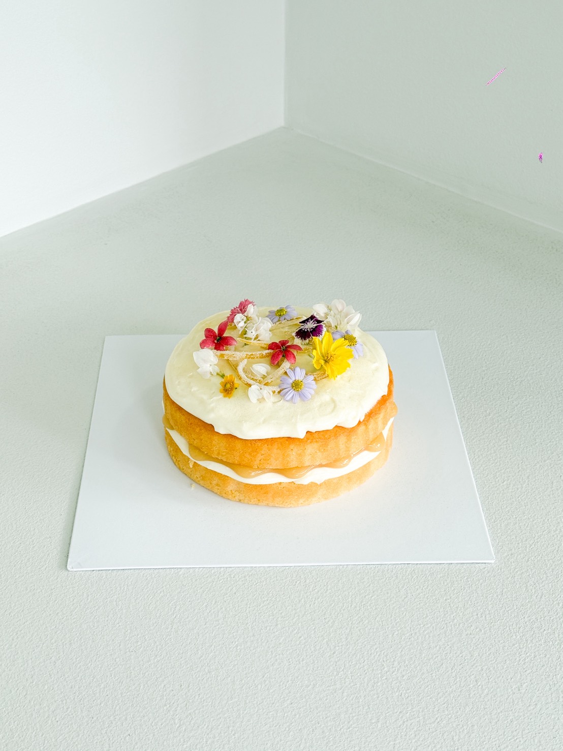 A two-layer vanilla cake with white icing, decorated with colourful edible flowers, sits on a white square board against a light grey background.
