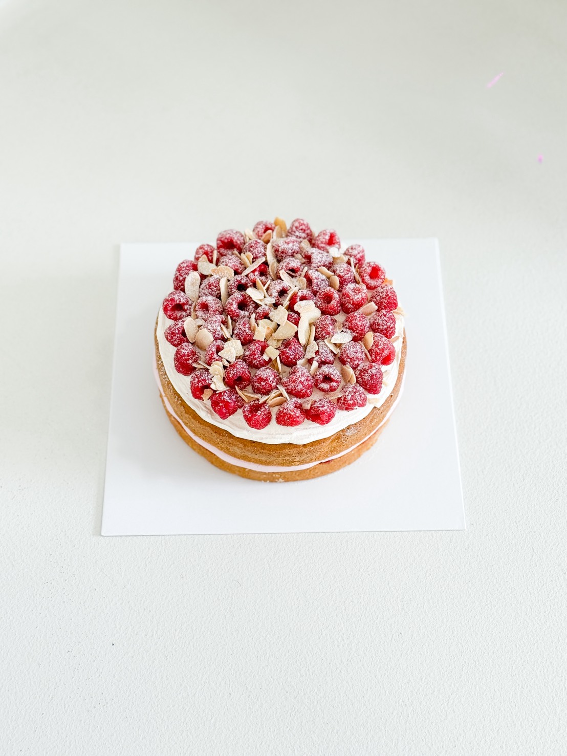 A round cake with white icing, topped with fresh raspberries, almond flakes, and icing sugar, sits on a white square base against a plain light background.