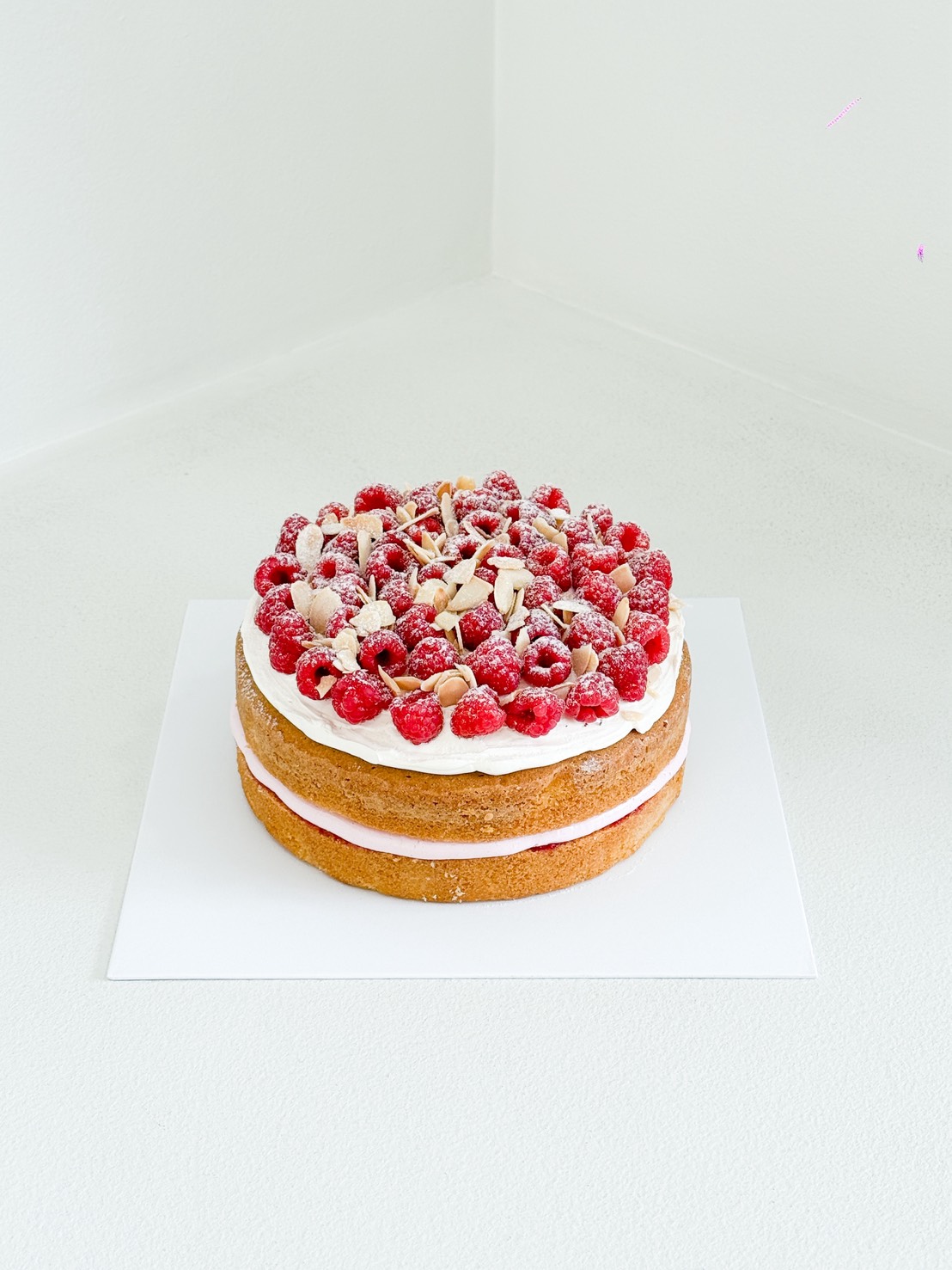 A two-layer sponge cake with cream and raspberry filling, topped with fresh raspberries, sliced almonds, and icing sugar, sits on a white square base in a minimalist white corner.