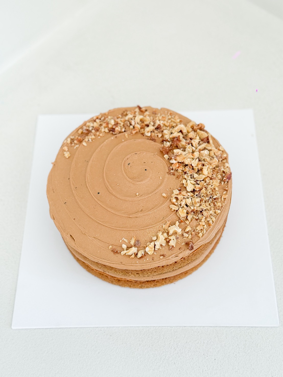 A round cake with light brown icing, decorated with swirled icing and a cluster of chopped nuts on one side, sits on a white square board against a light background.