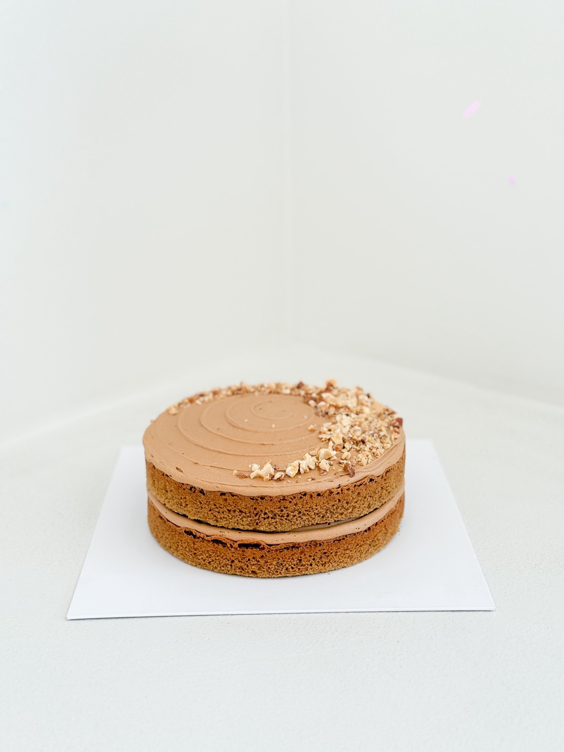A round, three-layer cake with light brown icing and a topping of chopped nuts on one side, displayed on a white square board against a plain white background.