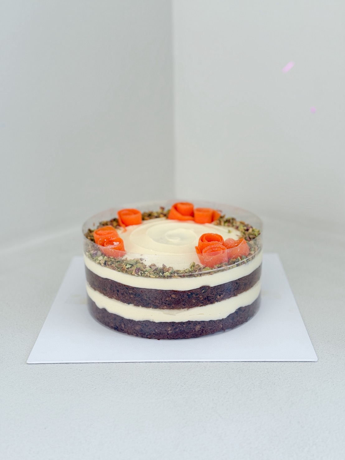 A round, layered carrot cake with white icing, topped with decorative orange swirls and a ring of chopped nuts, displayed on a white square surface against a plain background.