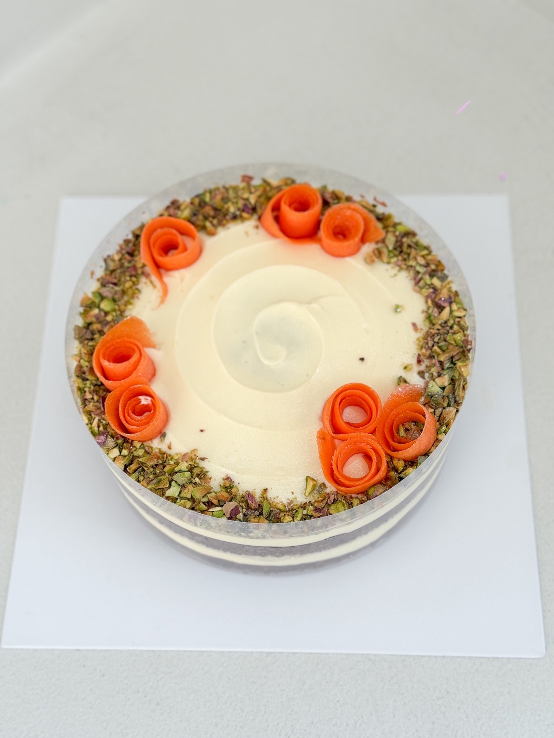 A round cake topped with white icing, decorated with crushed pistachios around the edge and orange carrot curls arranged on top, sitting on a white square board.