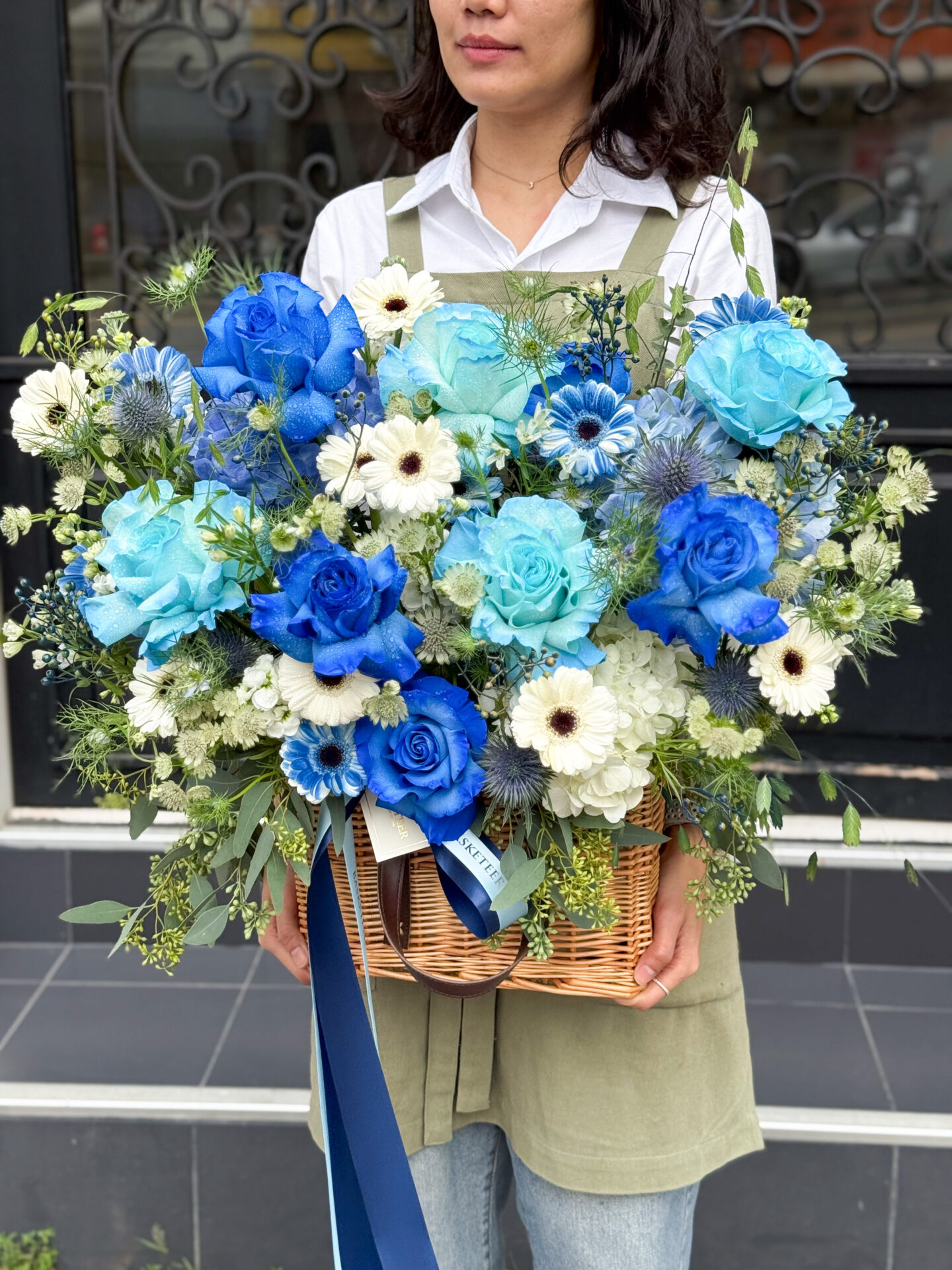 A person in a green apron holds a large wicker basket filled with blue and white roses, pink roses, white daisies, and greenery—reminiscent of a Carnations & Gerbera Basket Bouquet—with a blue ribbon cascading down the front.