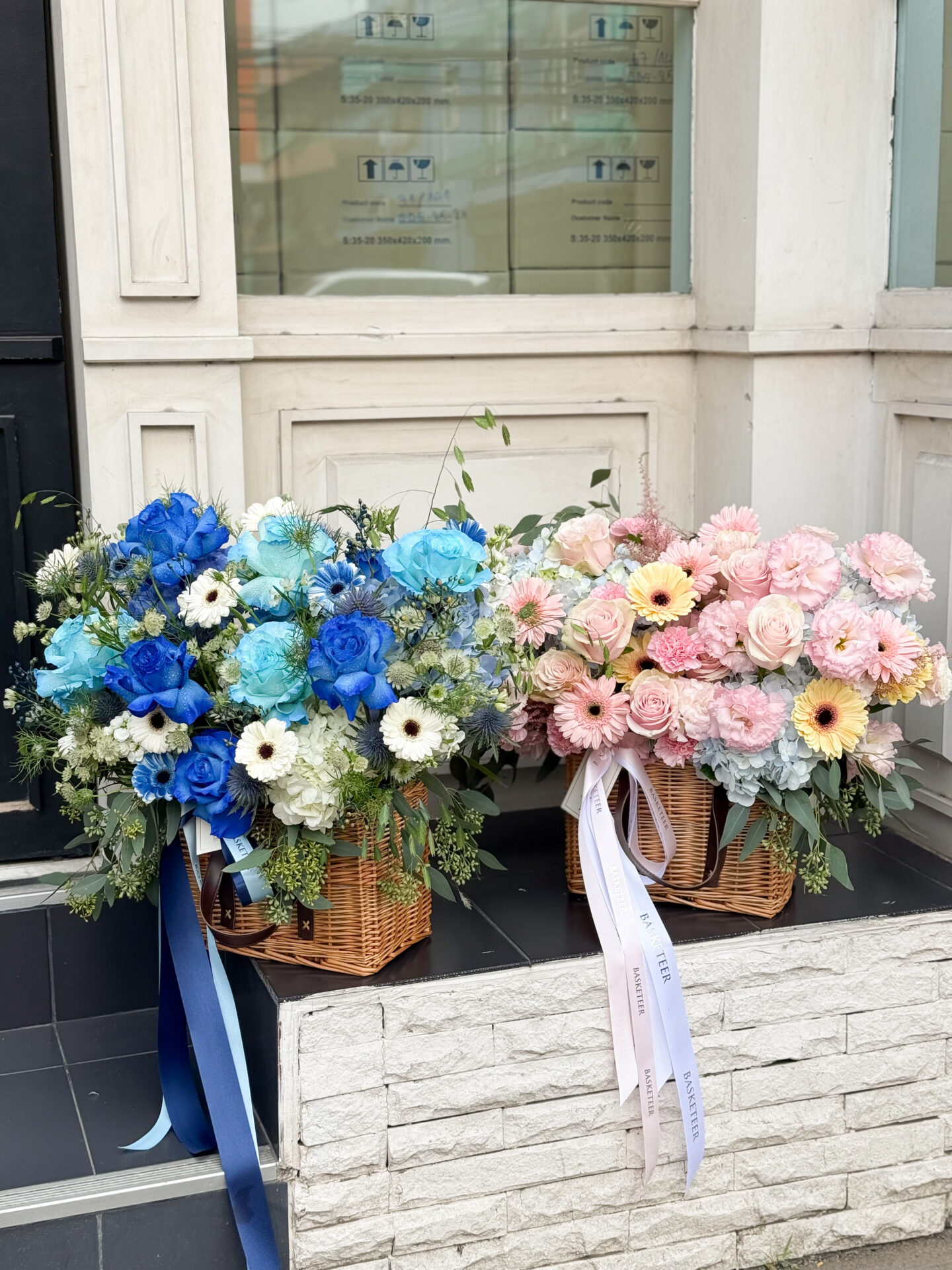 Two wicker baskets filled with flower arrangements: one with blue and white flowers including blue roses and daisies, and the other showcasing a Pink Roses, Carnations & Gerbera Basket Bouquet. Both have decorative ribbons and lush foliage.
