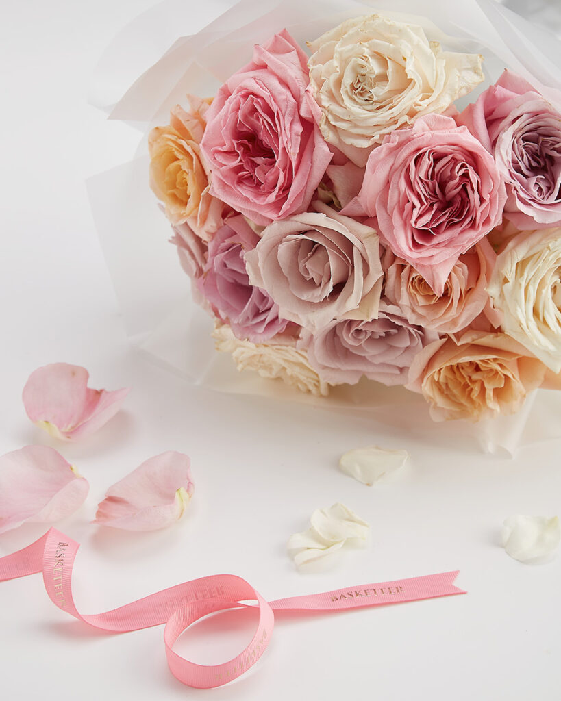A bouquet of pastel-coloured roses, including pink, peach, and cream hues, is wrapped in white paper. Scattered rose petals and a pink ribbon with “BASKETTEK” printed on it are placed on a white surface nearby.