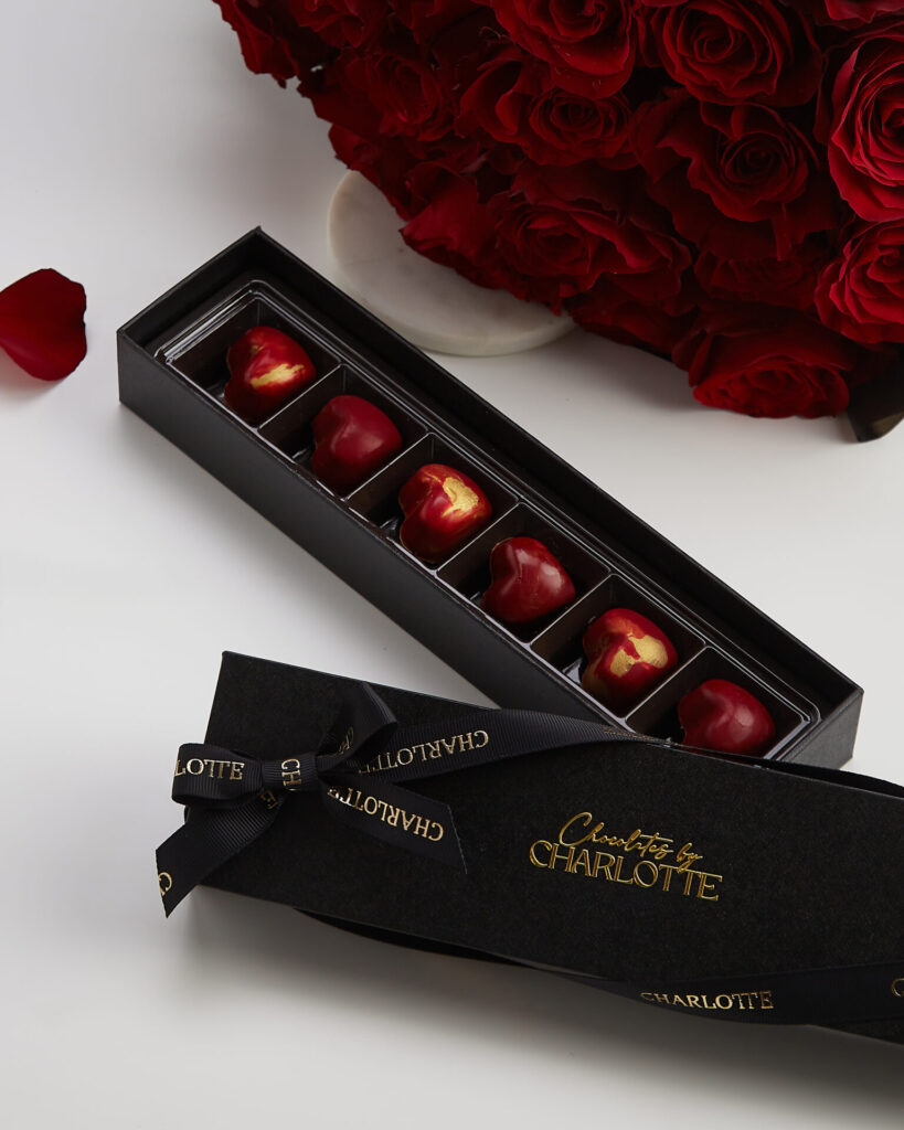 A black gift box labelled "CHARLOTTE" contains six red heart-shaped chocolates with gold accents, placed next to a bouquet of red roses on a white surface.