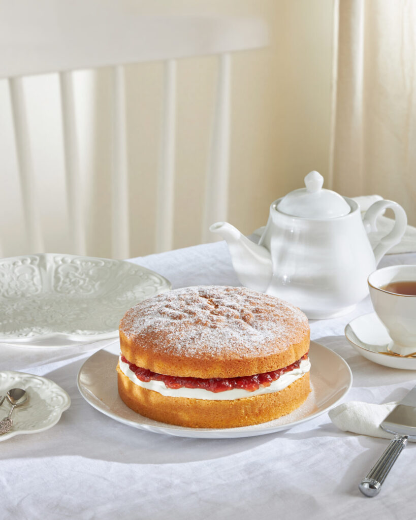 A traditional Victoria Sponge cake styled with a vintage teapot and recipe book, representing Grandma Amy's original 1940s British recipes used at Charlotte Bakery.