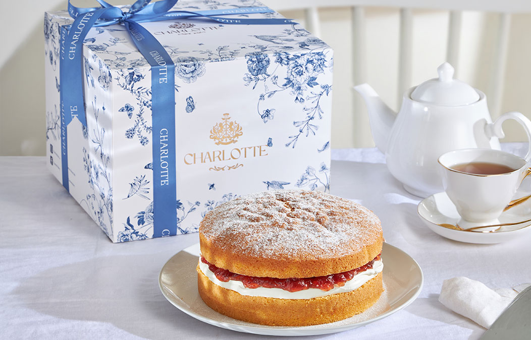 Victoria Sponge Cake on a plate with a white teapot and a Charlotte Bakery blue floral gift box in the background.