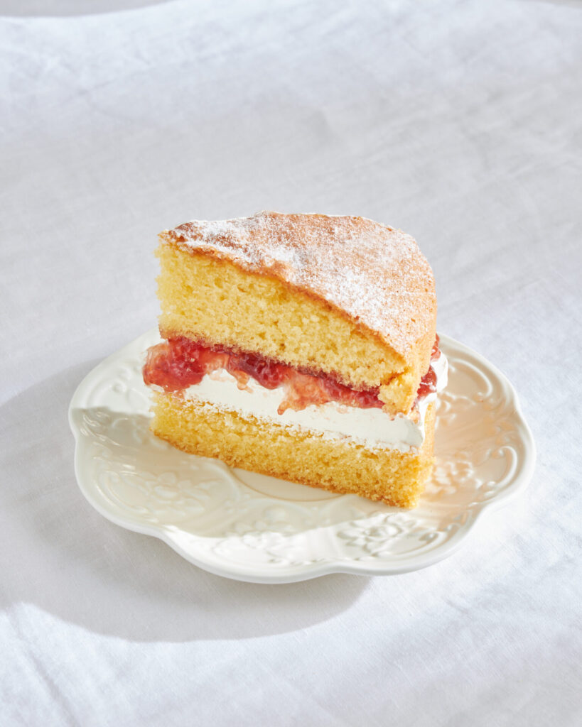 Close-up of a slice of Victoria Sponge cake on a white plate, showing the fluffy, buttery crumb texture.