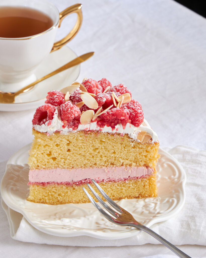 A slice of premium sponge cake topped with fresh cream and raspberries, plated with a gold spoon and tea cup.