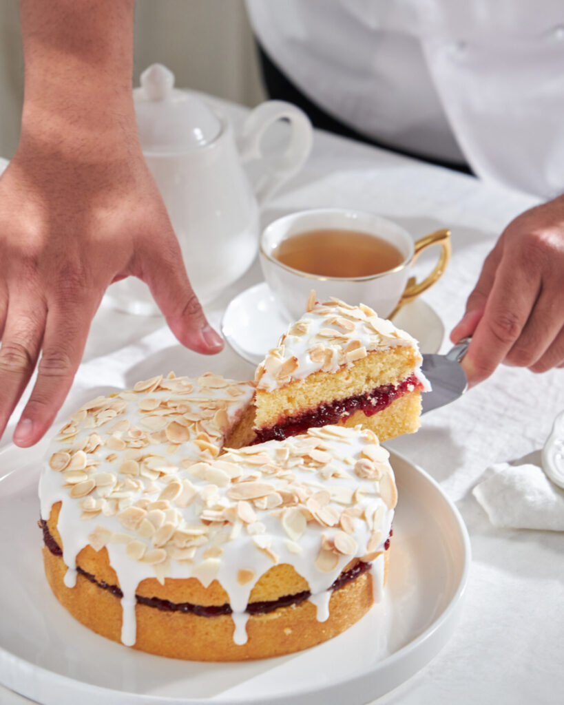 Slicing a glazed vanilla sponge cake, styled with a tea set to illustrate the perfect afternoon tea pairing.