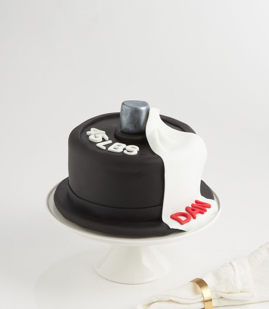 A black fondant cake on a white stand, decorated with a grey hammer, white and red letters spelling "DAD," and a white fondant drape. A folded serviette with a gold ring is beside the cake.