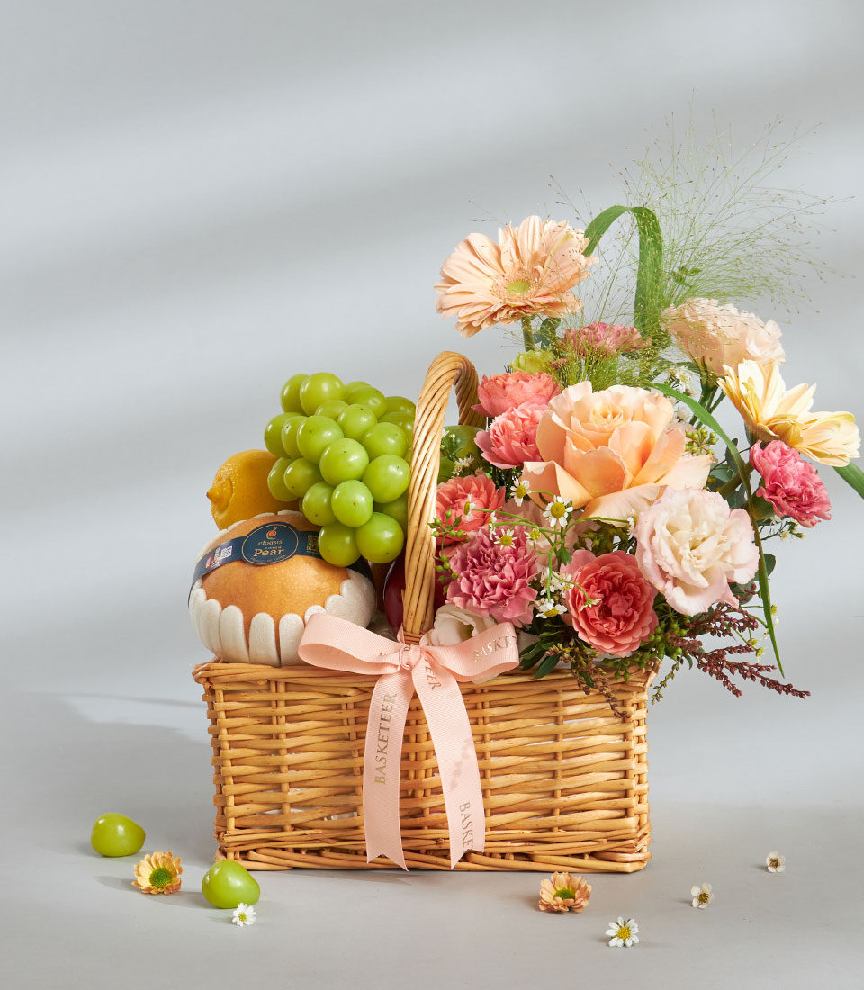 The Soft Peach Premium Fruit & Floral Basket features a wicker basket with a pink ribbon, holding green grapes, a pear, and peach and pink flowers—roses and carnations—against a light grey background, with scattered grapes and blooms nearby.