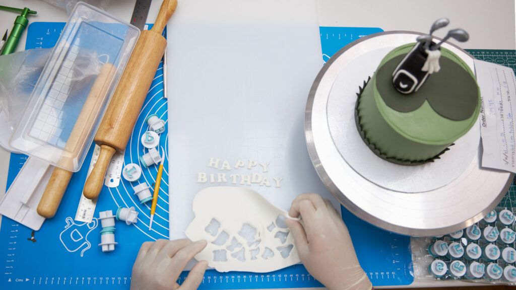 Hands preparing fondant “Happy Birthday” letters beside tools for a Charlotte Bakery custom cake.