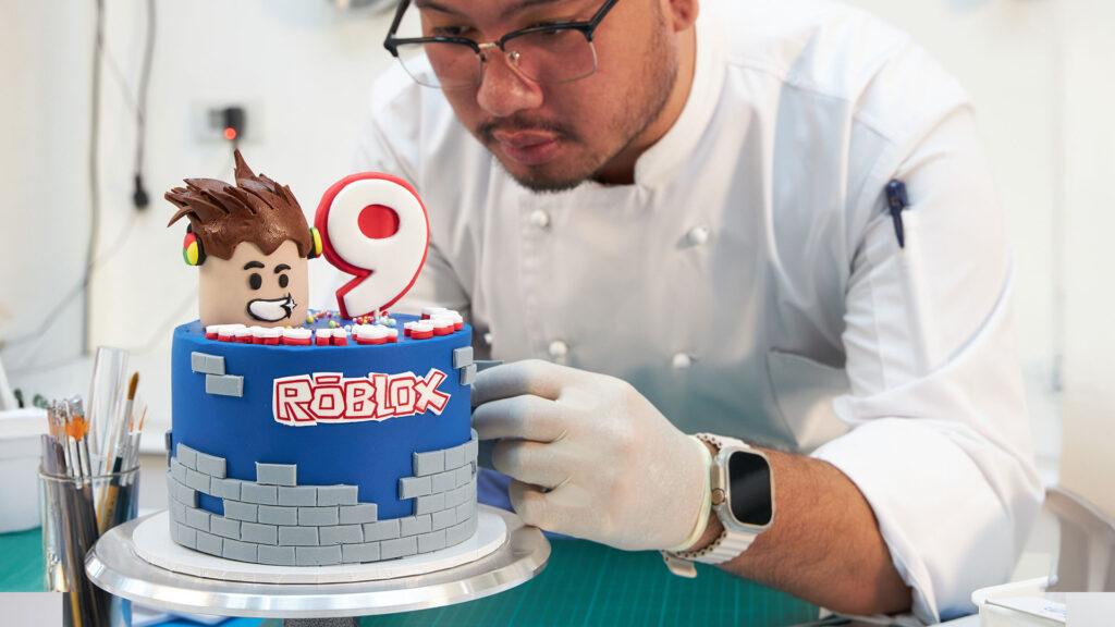 Charlotte Bakery chef decorating a Roblox-themed birthday cake with fondant details.