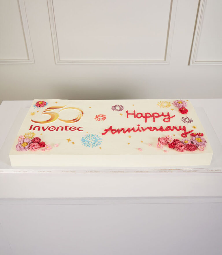 A rectangular cake with "50 Inventec" and "Happy Anniversary" written on it, decorated with colourful flowers and confetti designs, displayed on a white table against a white wall.