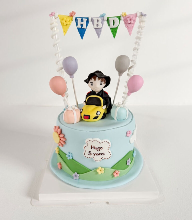 Fondant birthday cake with a child figure driving a toy car and pastel decorations by Charlotte Bakery in Bangkok.