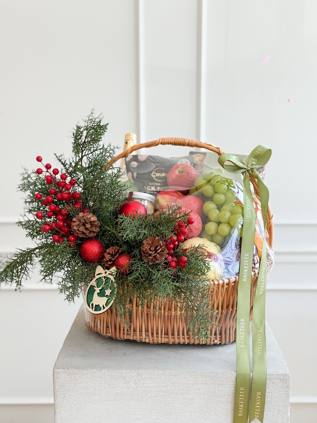 A wicker basket filled with apples, grapes, and snacks, decorated with pine cones, red berries, evergreen branches, red baubles, and a green ribbon, sitting on a grey pedestal against a white wall.