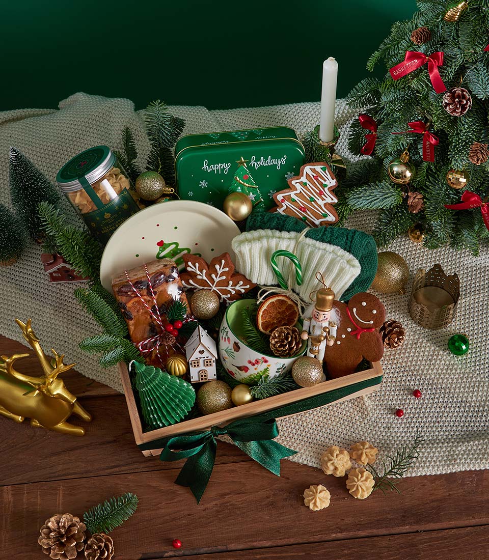 A festive holiday gift hamper filled with biscuits, a candle, baubles, a mug, a Christmas tree decoration, and treats, surrounded by fir branches, a gold reindeer, and holiday décor on a wooden floor.
