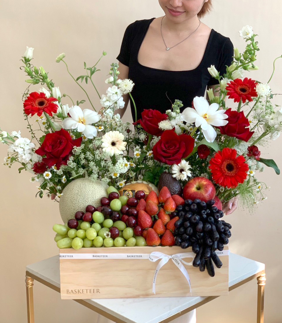 A woman in a black dress stands behind a wooden box filled with grapes, strawberries, apples, and melons, arranged with red and white flowers on a small table against a beige background.