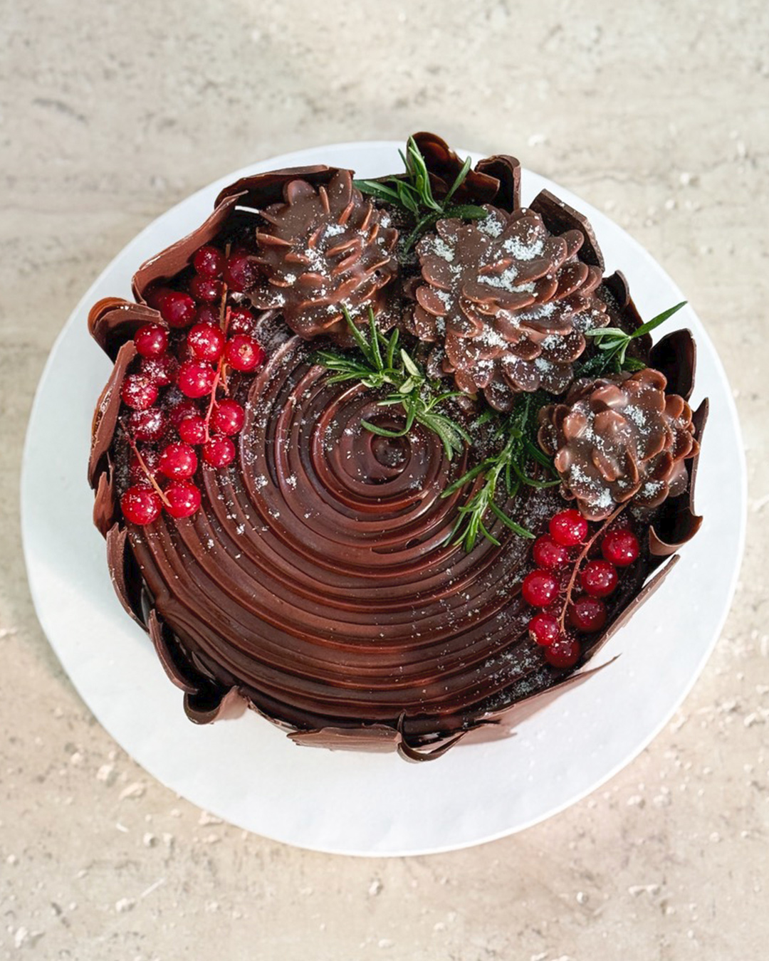 A Chocolate Ganache Christmas Cake, decorated with chocolate swirls, red berries, rosemary sprigs, and chocolate pinecones, is lightly dusted with icing sugar and sits on a white plate against a neutral background.