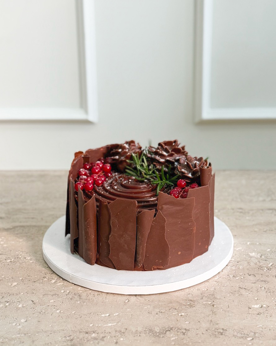 A Chocolate Ganache Christmas Cake decorated with chocolate shards, red berries, and rosemary sprigs sits on a white round cake board atop a beige surface, with a white panelled wall in the background.